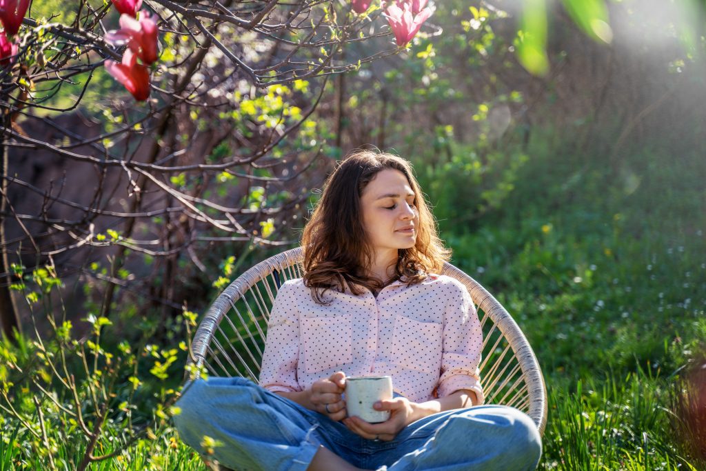 Young beautiful woman enjoying relaxing in the spring while sitting in the blooming garden of her home with a cup of tea