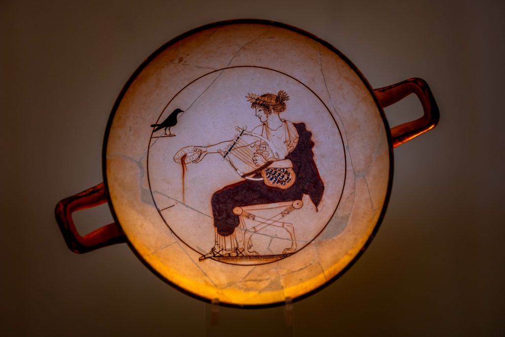 White ground kylix, found in a tomb of Delphi. Anonymous Athenian vase painter. Apollo is depicted crowned with a wreath of myrtle leaves, seated on a stool and dressed in white peplos and red himation draped over his left shoulder.