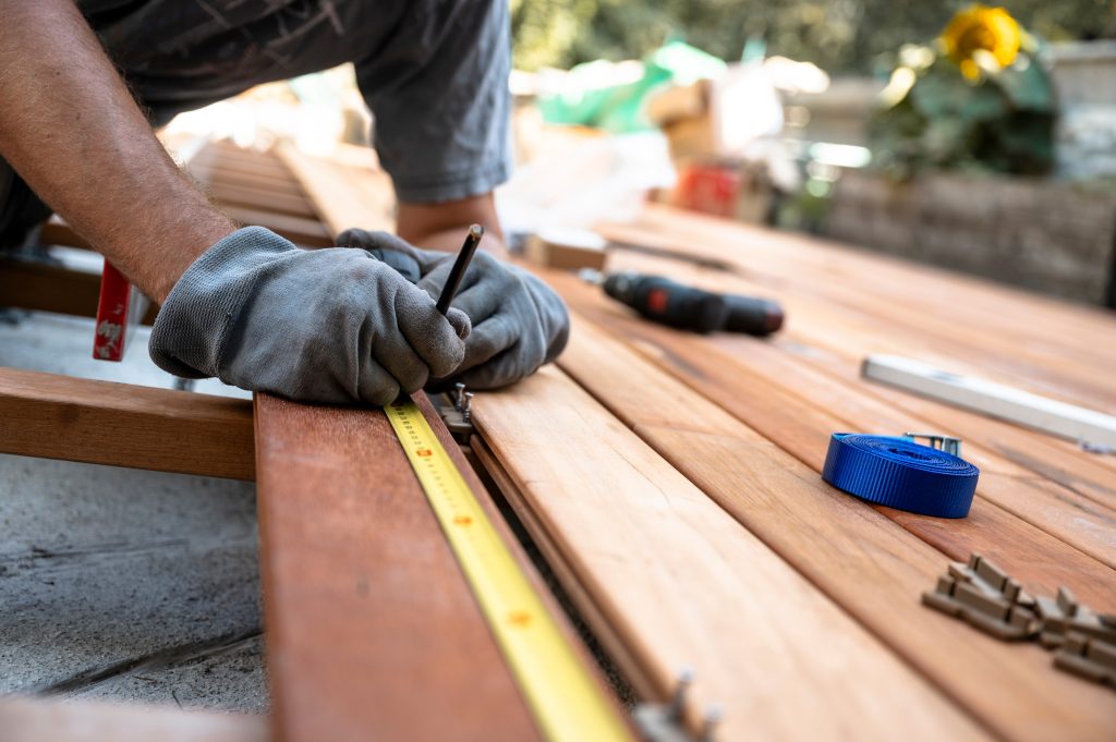 Low angle view of a worker with protective gloves marking a spot on wooden plank while placing it on a foundation for outside patio. Renovation and diy project.