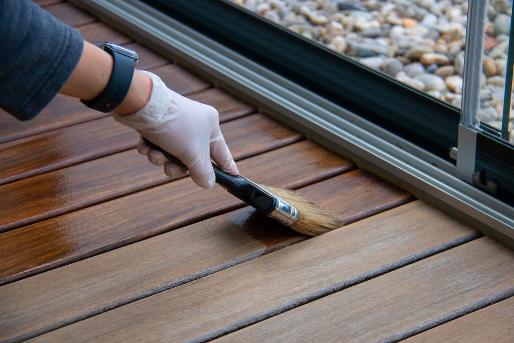 Deck staining, worker applying deck oil on decking boards with paint brush, hardwood terrace renovation