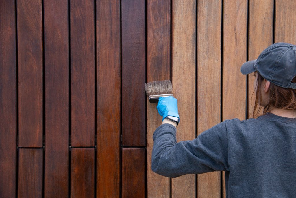 Handywoman applying protective varnish or paint with brush on wood fence wall cladding close up. Hardwood cladding house improvement diy concept