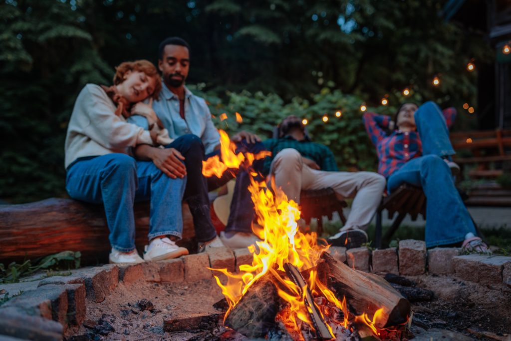 Close up shot of a fire burning in a fire pit with four people relaxing around it on a cozy summer evening in the background.