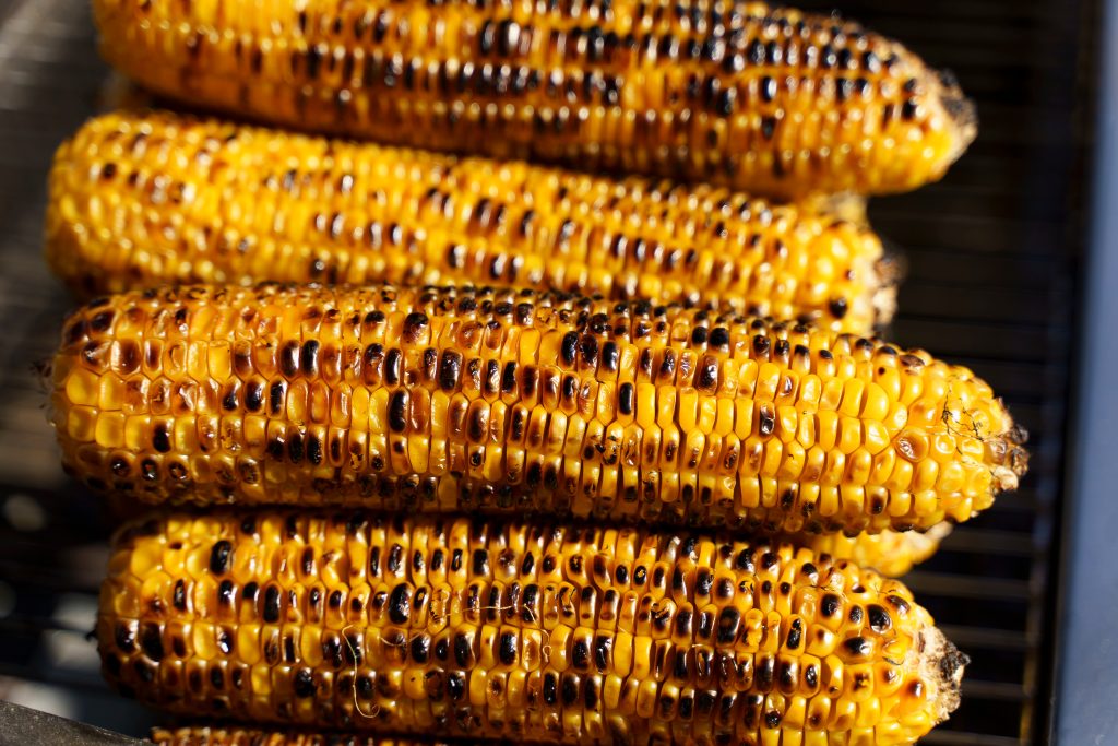 Close-up image of golden corn cobs being grilled over an open flame. The charred texture and glowing coals evoke a sense of summer, outdoor cooking, and mouthwatering street food traditions. Perfect for culinary content, food blogs, barbecue themes, picnic promotions, and summer festival visuals.