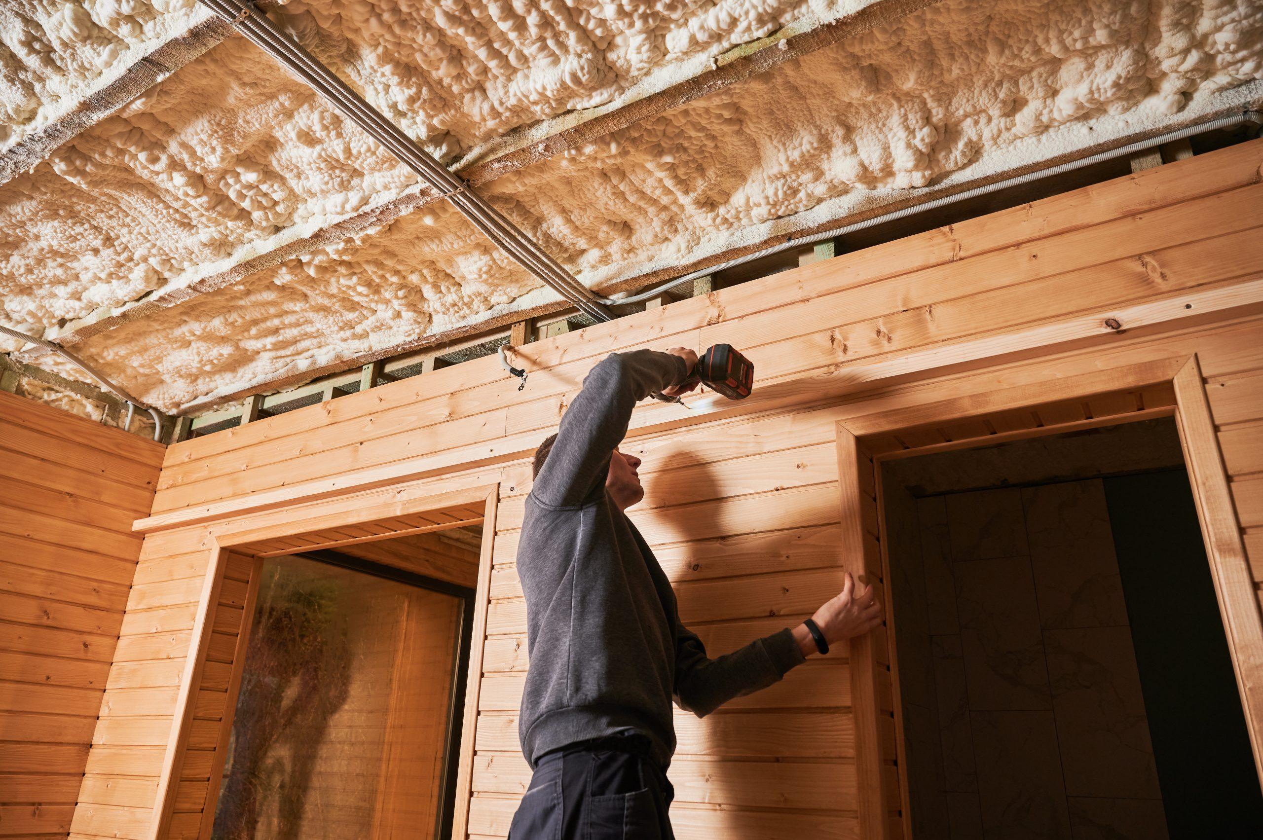 Man fixing wooden trim above doorway with cordless drill during installation work. Indoor construction with timber cladding, window opening, insulation and wiring, home renovation concept