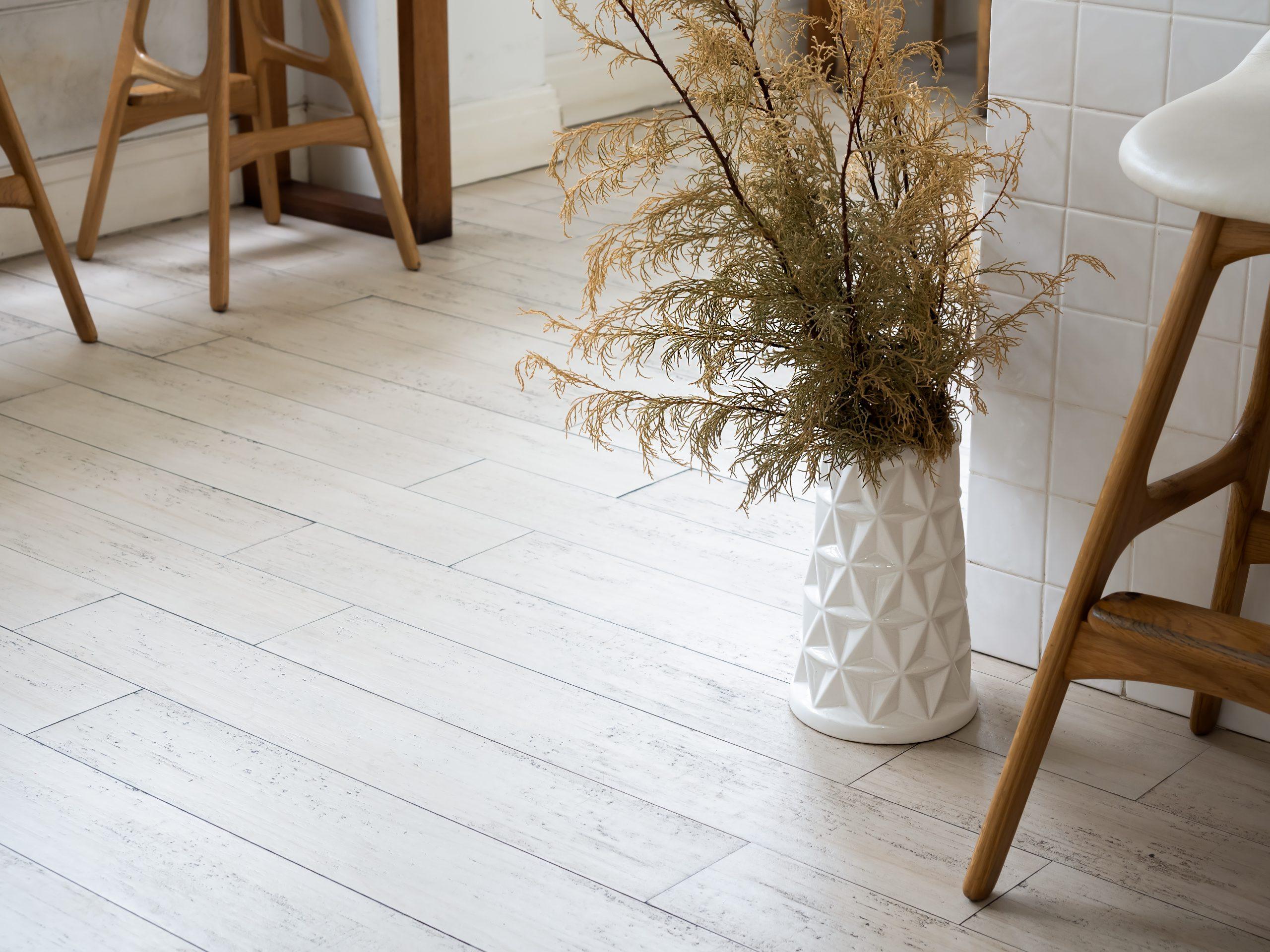 Timber Flooring Guide – Choosing the Right Floor for Your Home 5 Green leaves plant in modern white tall ceramic vase decoration on wood pattern rubber tiles near vintage wooden bar stool chair and white wall background with space. Home interior rustic brown tone.